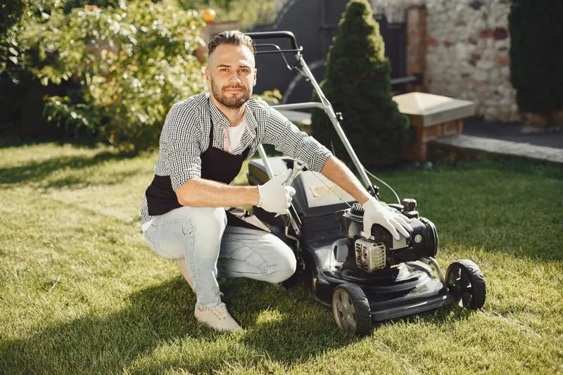 Adult man wearing apron and gloves maintains a lawn mower on a sunny day