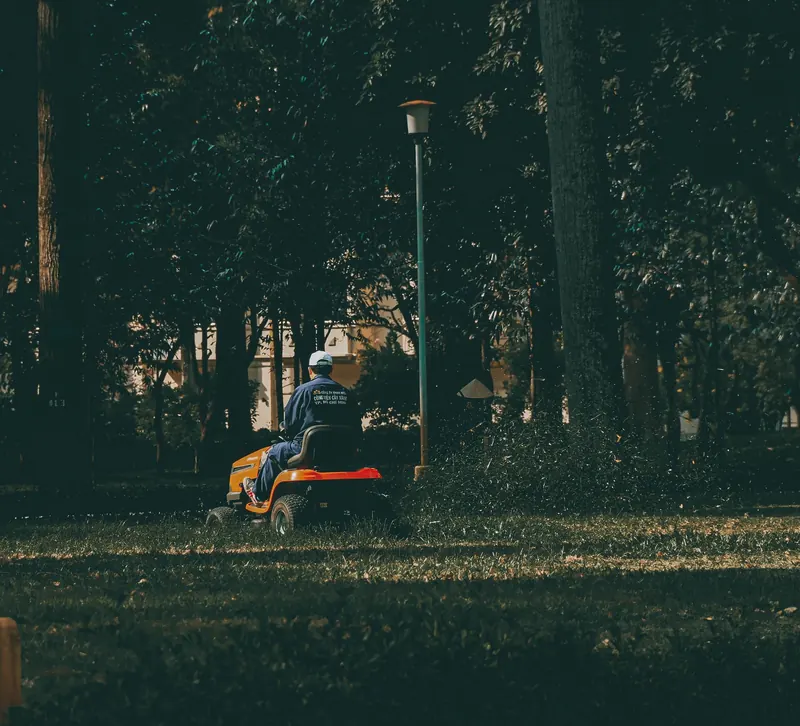 A man in coveralls operates a lawn mower in a verdant park setting, surrounded by lush trees and grass